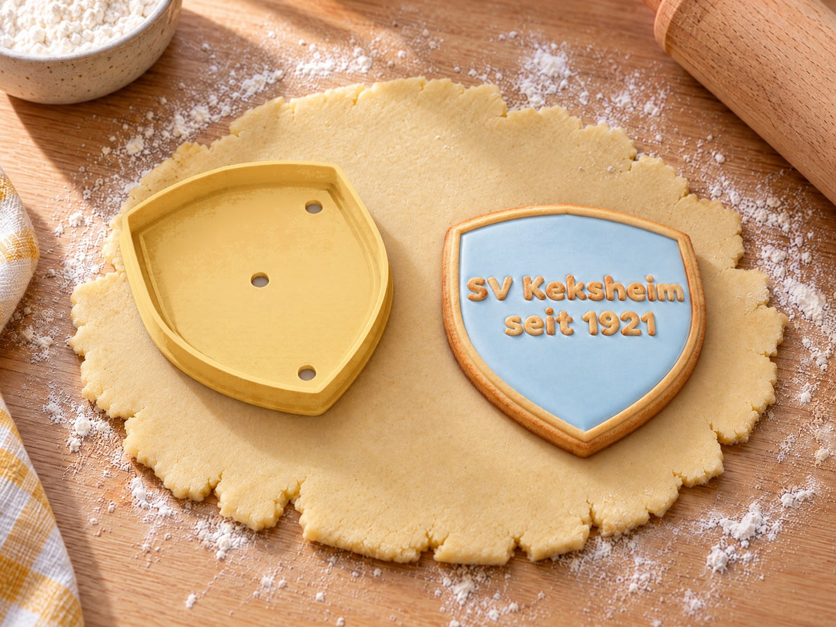Butter-gelber Wappenschild-Ausstecher neben gebackenem Schildkeks mit hellblauem Zuckerguss und geprägtem Text „SV Keksheim seit 1921“ auf ausgerolltem Teig und hellem Holz