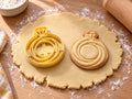 Butter-gelber Verlobungsring-Ausstecher mit Prägung neben Keks mit geschwungenem Ring und kleinem Solitär auf hellem Holz