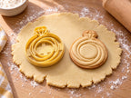 Butter-gelber Verlobungsring-Ausstecher mit Prägung neben Keks mit geschwungenem Ring und kleinem Solitär auf hellem Holz