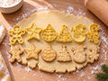 Set Weihnachts-Klassiker: 6 butter-gelbe Ausstecher mit Prägung nebeneinander mit gebackenen, geprägten Keksen auf ausgerolltem Teig und hellem Holz