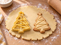 Butter-gelber Origami-Tannenbaum-Ausstecher mit Prägung neben gebackenem Keks mit gefalteten Dreiecksflächen und Stern auf hellem Holz