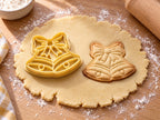 Butter-gelber Weihnachtsglocken-Ausstecher mit Prägung neben gebackenem Keks mit Schleife, Zierringen und Klöppeln auf hellem Holz