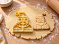 Butter-gelber Weihnachtsmann-Ausstecher mit Prägung neben gebackenem Keks mit Bommelmütze, Bart und Knopfmantel auf hellem Holz