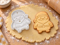 Weihnachtsmann-Ausstecher mit Prägung neben gebackenem Keks mit Mütze, Rauschebart, Gürtel und Stiefeln