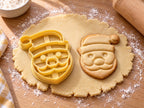 Butter-gelber Weihnachtsmann-Ausstecher mit Prägung neben gebackenem Keks mit großen Kulleraugen und Bommelmütze auf hellem Holz