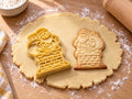 Butter-gelber Weihnachtsmann-Ausstecher mit Prägung neben gebackenem Keks mit Backsteinkamin und Geschenkesack auf hellem Holz