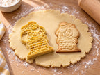 Butter-gelber Weihnachtsmann-Ausstecher mit Prägung neben gebackenem Keks mit Backsteinkamin und Geschenkesack auf hellem Holz