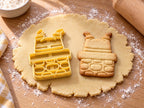 Butter-gelber Ausstecher mit Prägung neben gebackenem Keks: Weihnachtsmann-Stiefel ragen kopfüber aus einem Steinkamin, auf hellem Holz