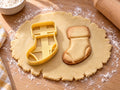 Butter-gelber Socken-Ausstecher mit Prägung neben gebackenem Keks mit breitem Umschlag, Ferse und Spitze auf hellem Holz