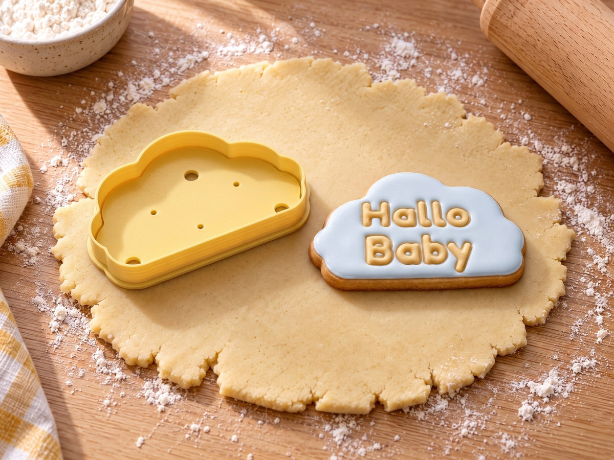 Butter-gelber Wolken-Ausstecher neben gebackenem Wolkenkeks mit hellblauem Zuckerguss und geprägtem Text „Hallo Baby“ auf ausgerolltem Teig und hellem Holz