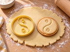 Butter-gelber Yin-und-Yang-Ausstecher mit Prägung neben gebackenem Keks mit S-Linie, zwei Punkten und Ring auf hellem Holz