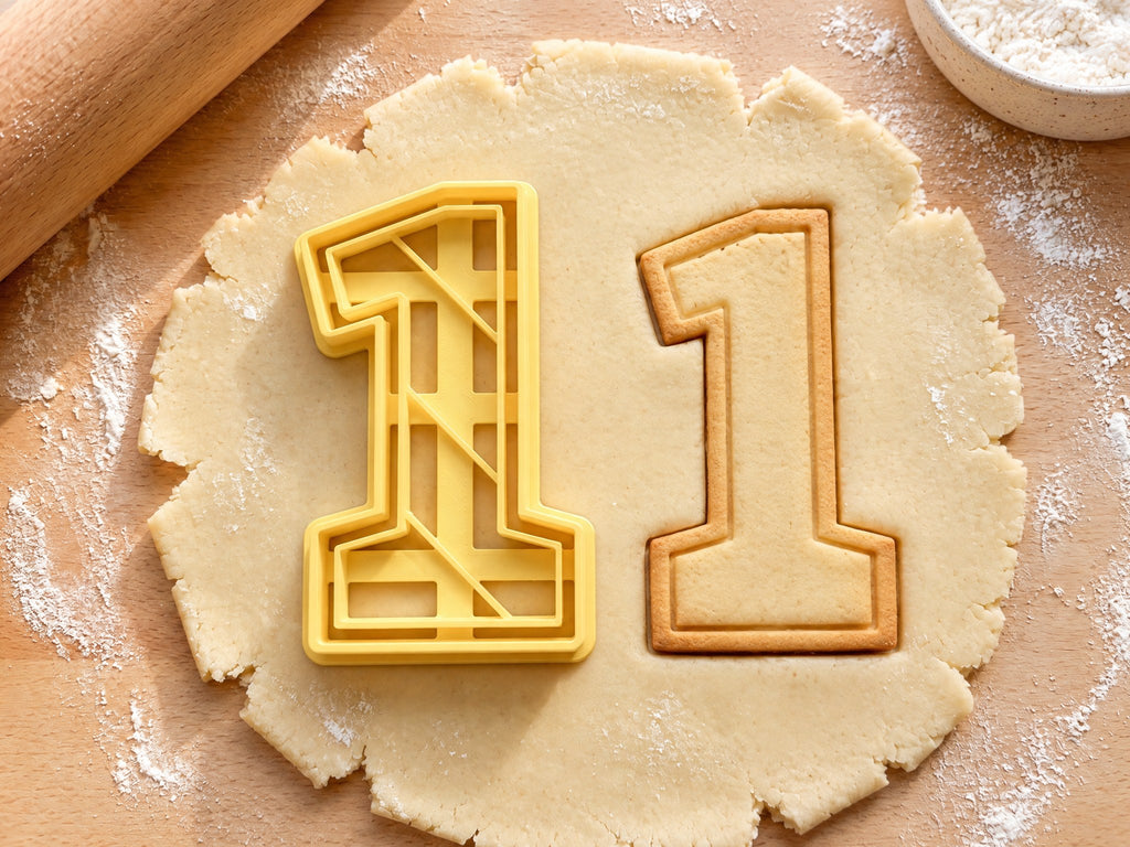 Butter-gelber Ausstecher Zahl 1 mit Prägung neben Keks in Form einer Eins mit Sockel und Konturlinie auf hellem Holz