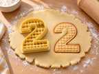 Butter-gelber Ausstecher Zahl 2 mit Prägung neben gebackenem Keks mit feinem Gittermuster auf hellem Holz