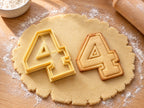 Butter-gelber Ausstecher Zahl 4 mit Prägung neben Keks in Form einer geschlossenen Vier mit doppelter Kontur auf hellem Holz