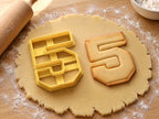 Butter-gelber Ausstecher Zahl 5 mit Prägung neben Keks in Form einer kantigen Fünf mit Konturlinie auf hellem Holz