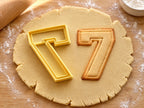 Butter-gelber Ausstecher Zahl 7 mit Prägung neben Keks in Form einer kantigen Sieben mit doppelter Kontur auf hellem Holz