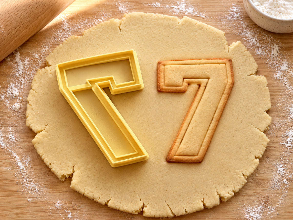 Butter-gelber Ausstecher Zahl 7 mit Prägung neben Keks in Form einer kantigen Sieben mit doppelter Kontur auf hellem Holz