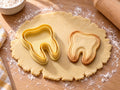 Butter-gelber Zahn-Ausstecher mit Prägung als schlichter Umriss neben gebackenem Keks mit feiner Randlinie auf hellem Holz