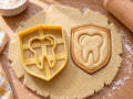 Butter-gelber Ausstecher mit Prägung als Zahn im Wappenschild neben gebackenem Keks mit Zahn und Schildrand auf hellem Holz