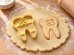 Butter-gelber Ausstecher mit Prägung als Zahn mit Pflaster neben gebackenem Keks mit zwei gekreuzten Pflastern auf hellem Holz
