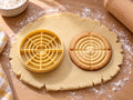 Butter-gelber Zielscheiben-Ausstecher mit Prägung neben gebackenem Keks mit vielen Ringen und feinem Fadenkreuz auf hellem Holz