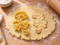 Butter-gelber Zuckerstangen-Ausstecher mit Prägung neben gebackenem Keks mit breiten Schrägstreifen und Schleife auf hellem Holz